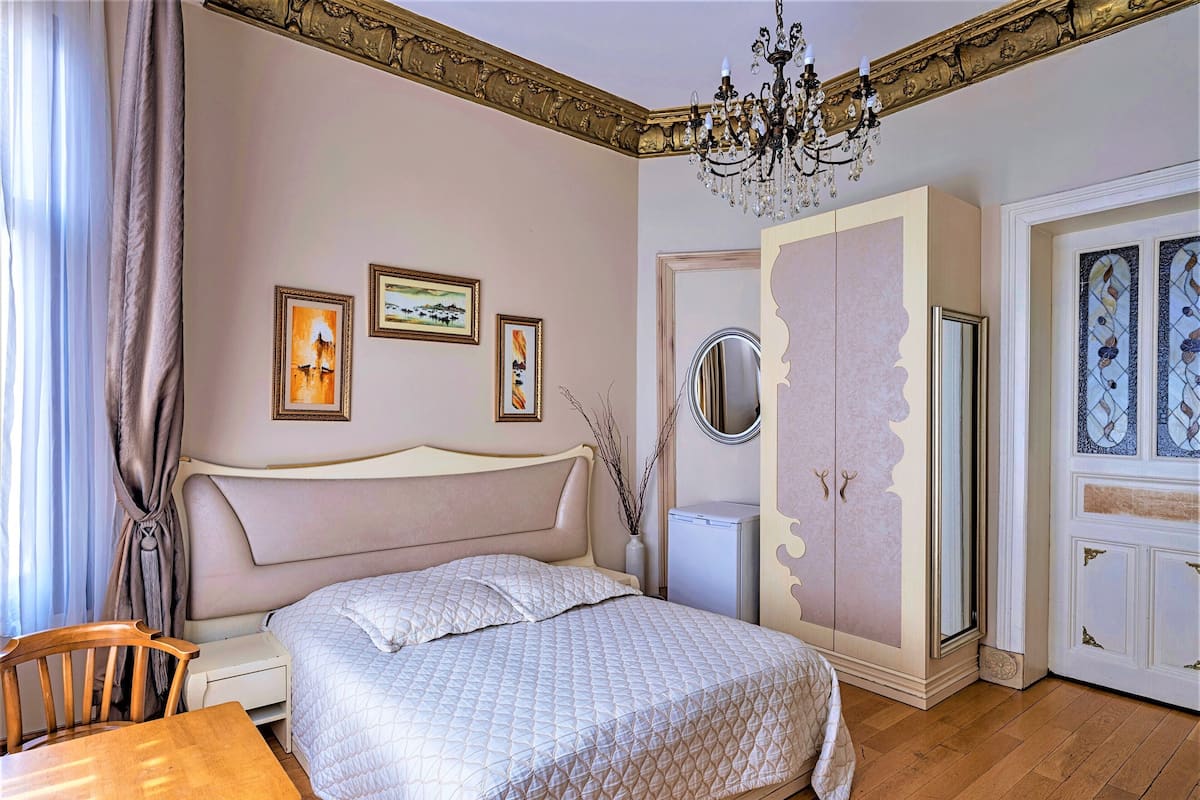 A bedroom with a bed, a wooden chair, a table, a chandelier, and a wardrobe.
