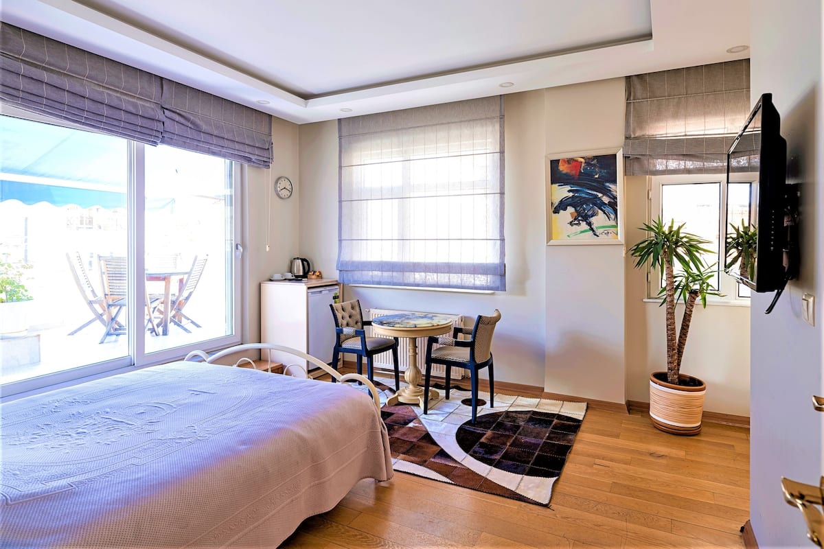 A bedroom with a bed, a small desk, a chair, a potted plant, and a window with blinds.
