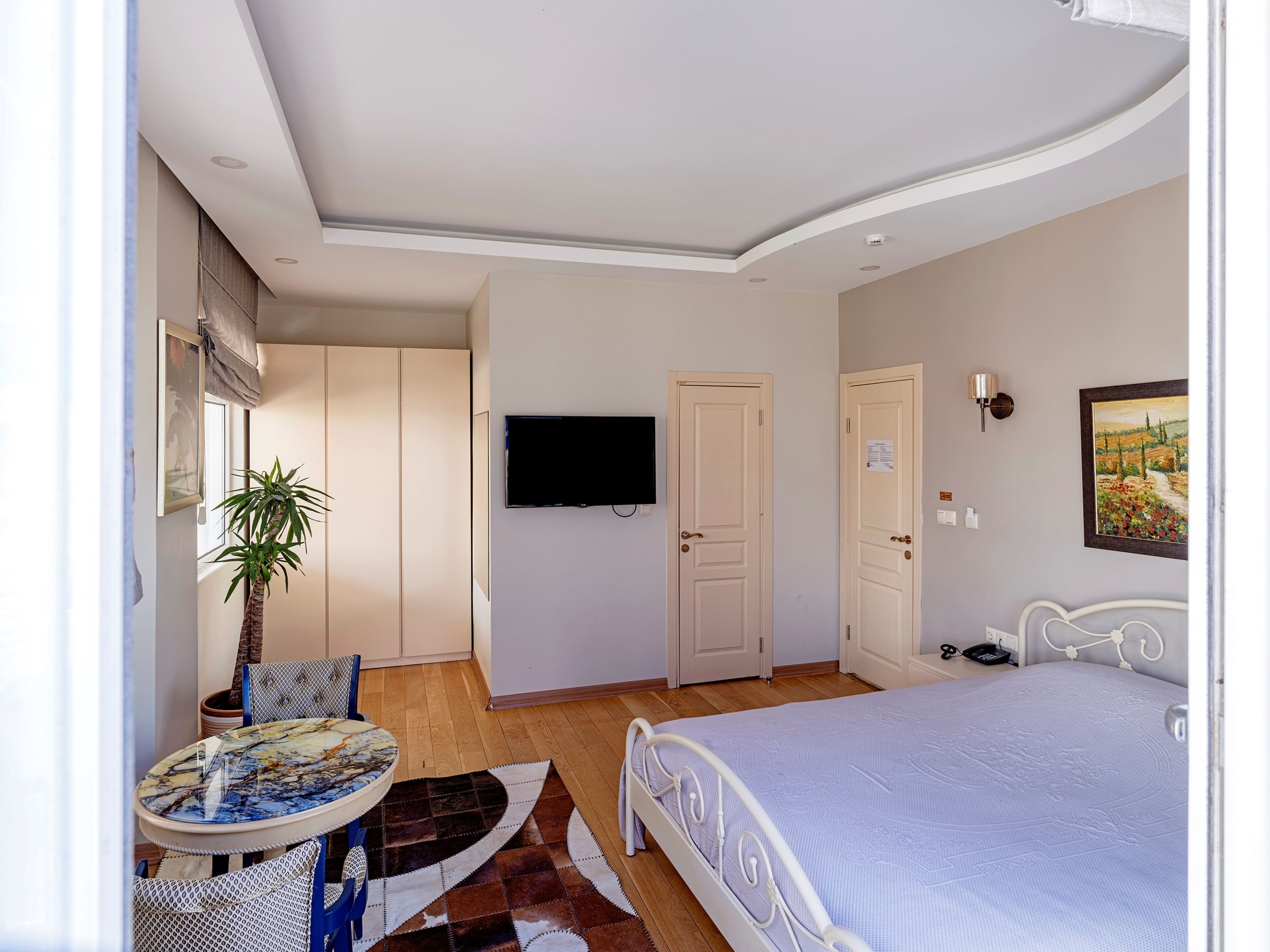 A bedroom with a bed, a TV mounted on the wall, a desk with a chair, and a potted plant.