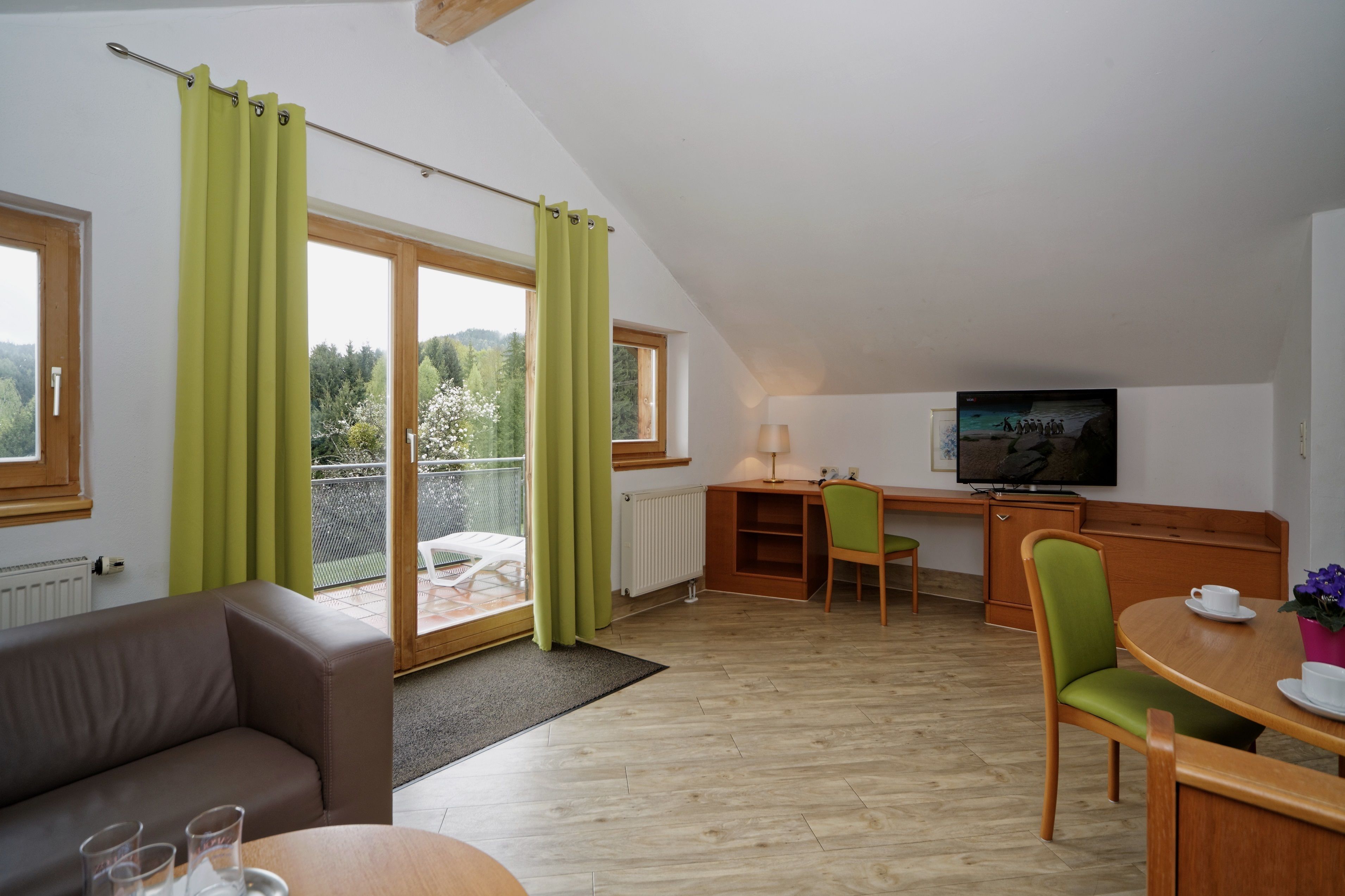 Ein modernes Zimmer mit Sofa, Esstisch und Fernseher. Es verfügt über einen Balkon mit Vorhängen und Blick ins Grüne.