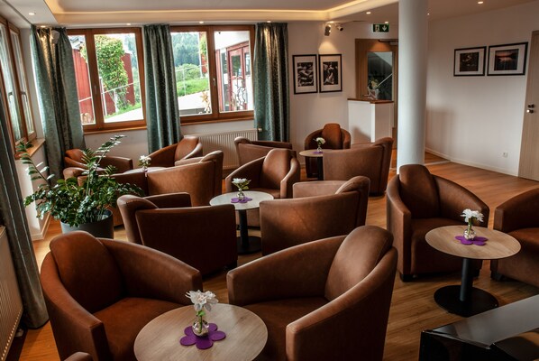A hotel lobby with brown leather chairs, small round tables with vases, large windows with curtains, and framed pictures on the wall.