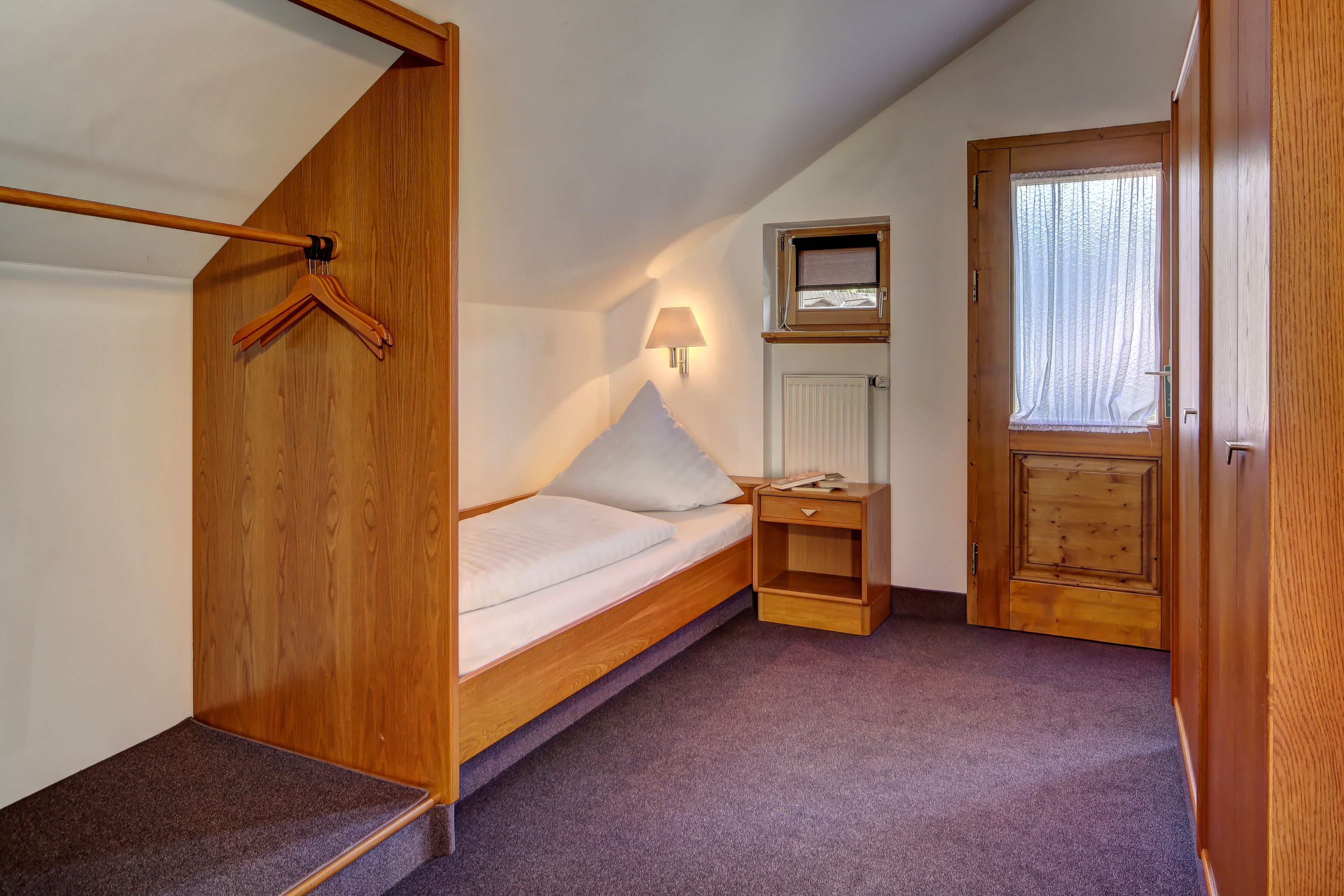 A small, cozy bedroom with a wooden bunk bed, a bedside table with a lamp, a small shelf, and a window with curtains.