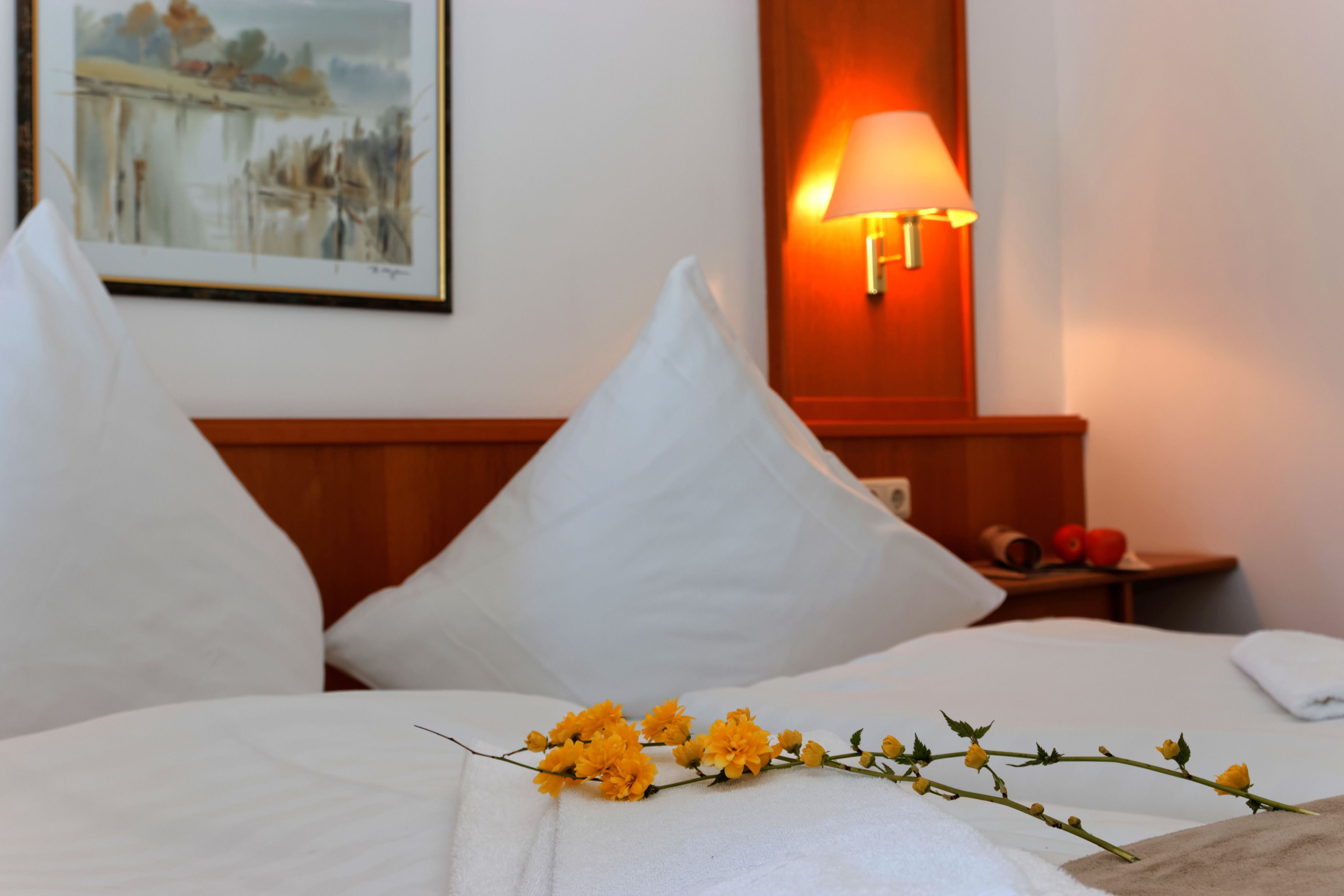 Cama bem arrumada com lençóis brancos, um arranjo floral sobre a cama e um quadro na parede.