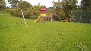 Children can play on the playground's slide and climbing elements, surrounded by trees and a wooden swing set.