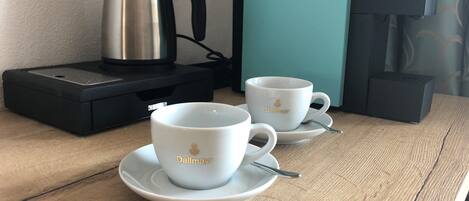 Two white cups with gold text on a wooden table, a black coffee maker, and a teal-colored box.