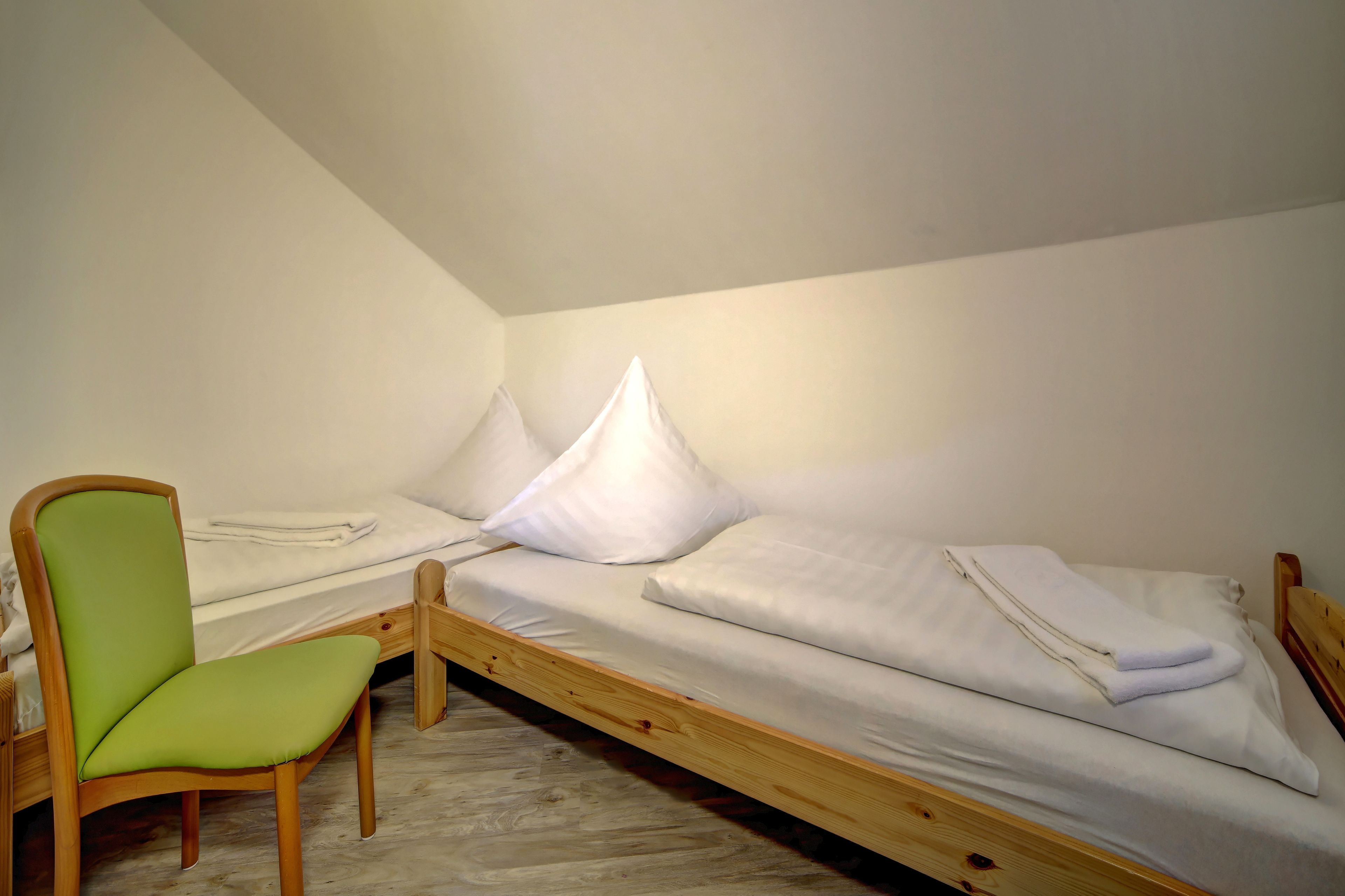 A small, single-room accommodation with a wooden bed, a green chair, and a white pillow.