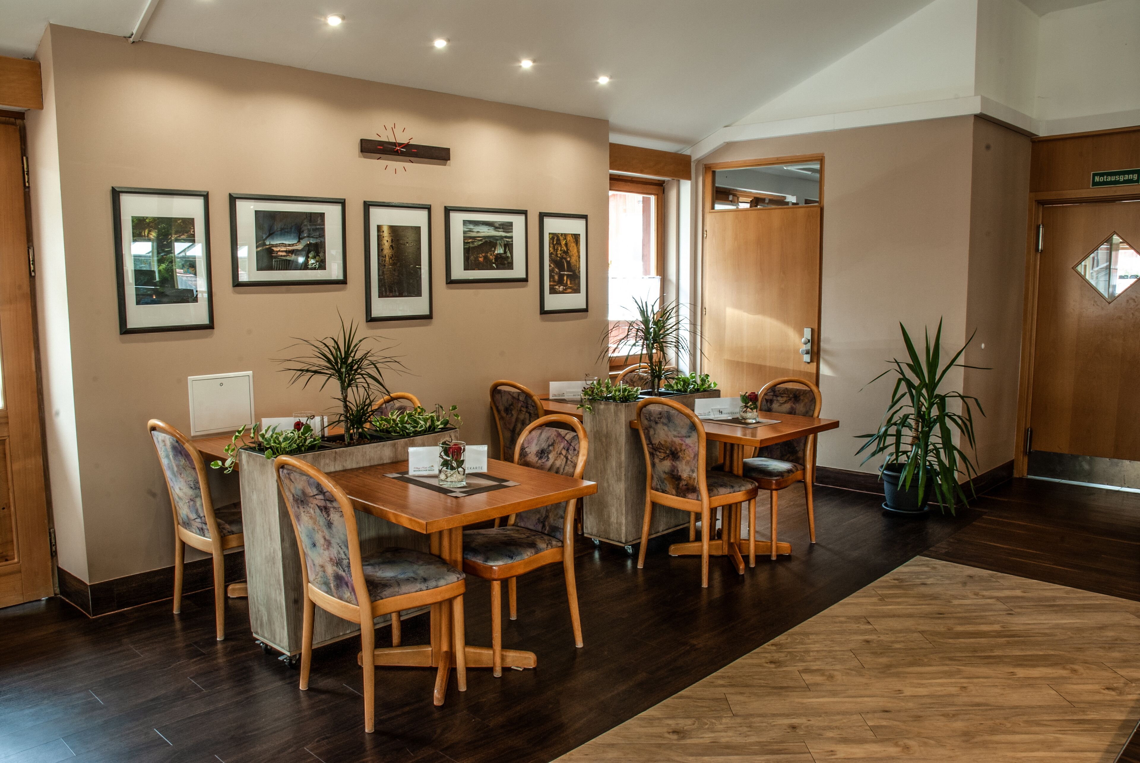 A dining area with wooden tables and chairs, framed pictures on the wall, and potted plants.