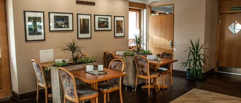 A dining area with wooden tables and chairs, framed pictures on the wall, and potted plants.