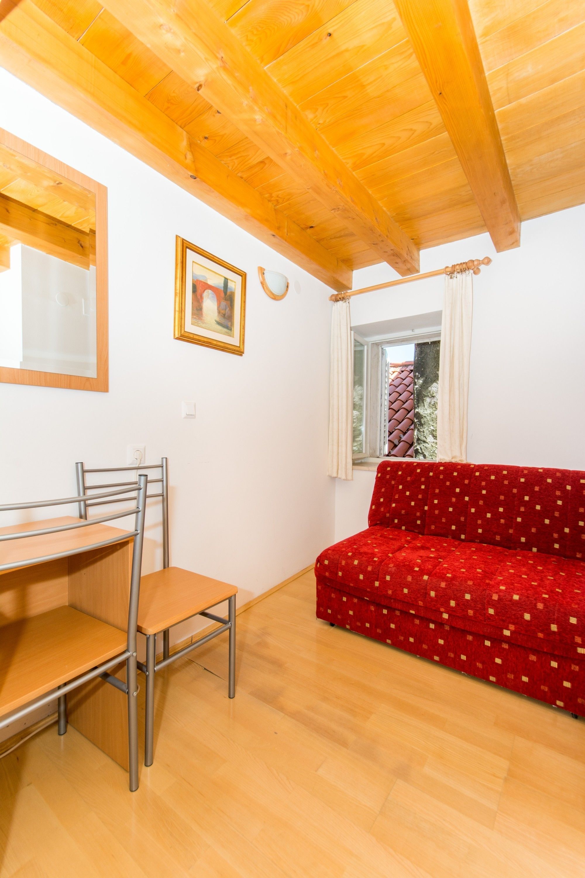Una habitación con techo de madera, un sofá rojo, una mesa de madera con una silla y una ventana con cortinas.