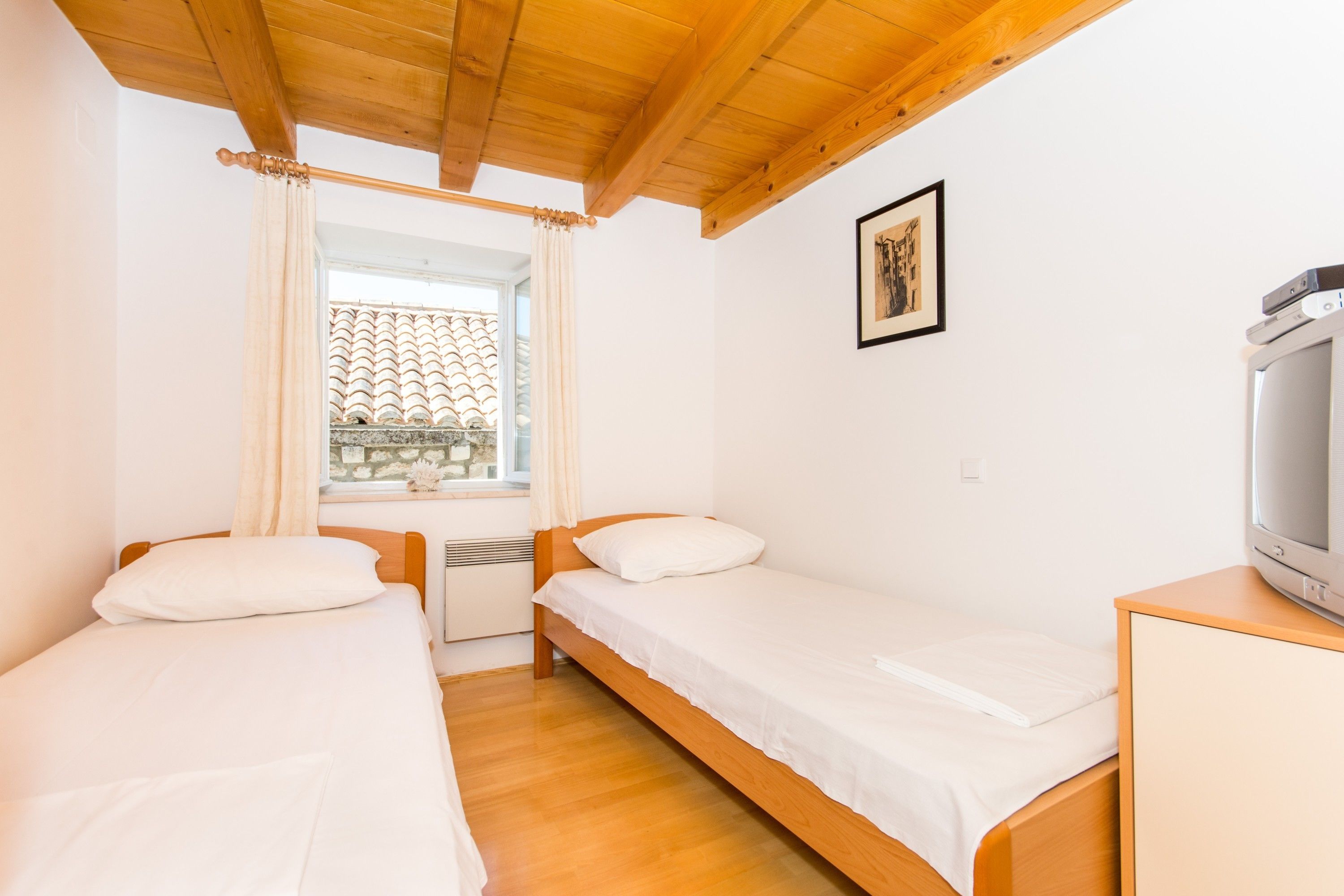 Una stanza piccola e pulita con due letti singoli, un soffitto in legno, una finestra vista su un tetto in tegole e una televisione su un mobile in legno.