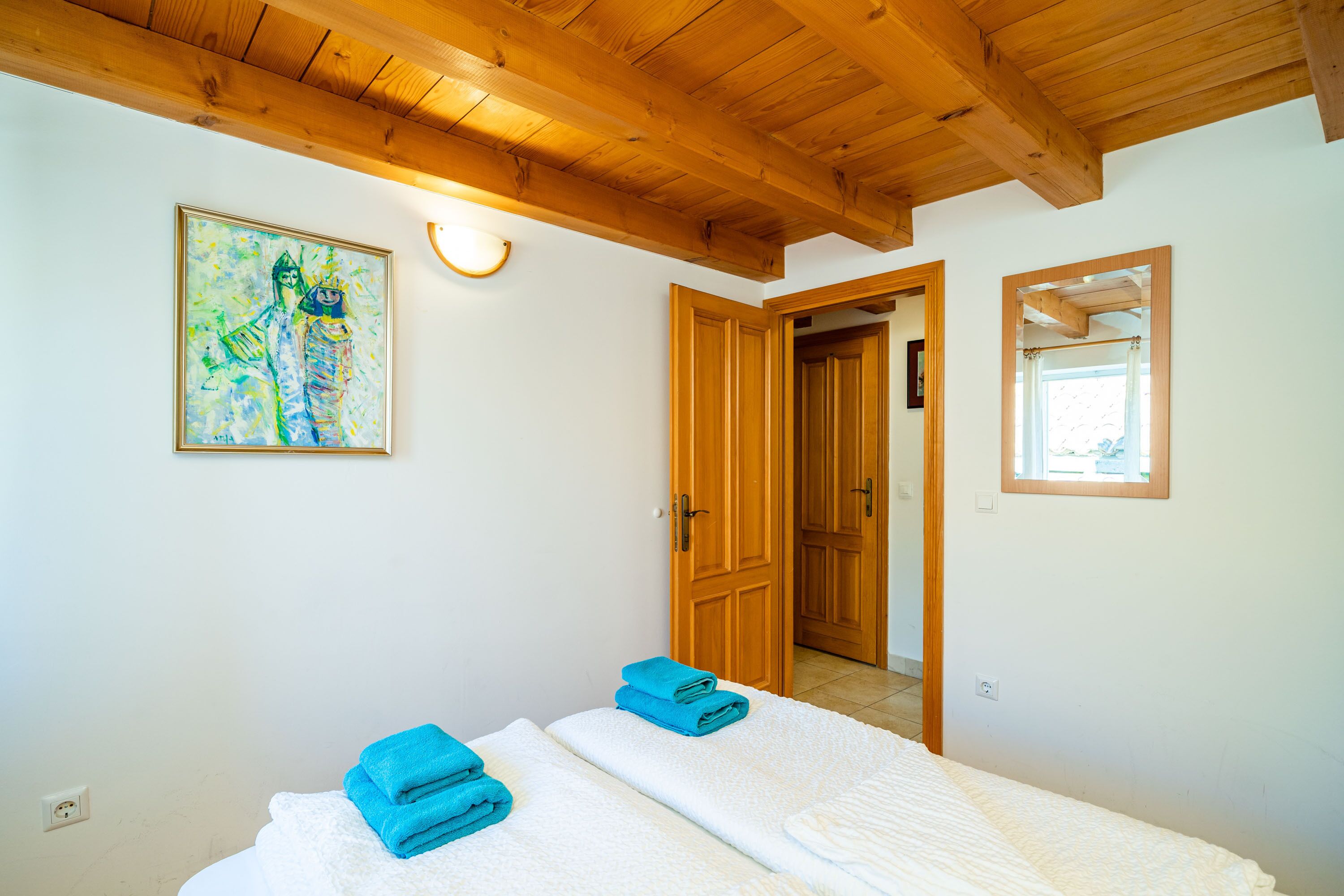 A room with two beds, wooden ceiling beams, a painting, and a door leading to another room.