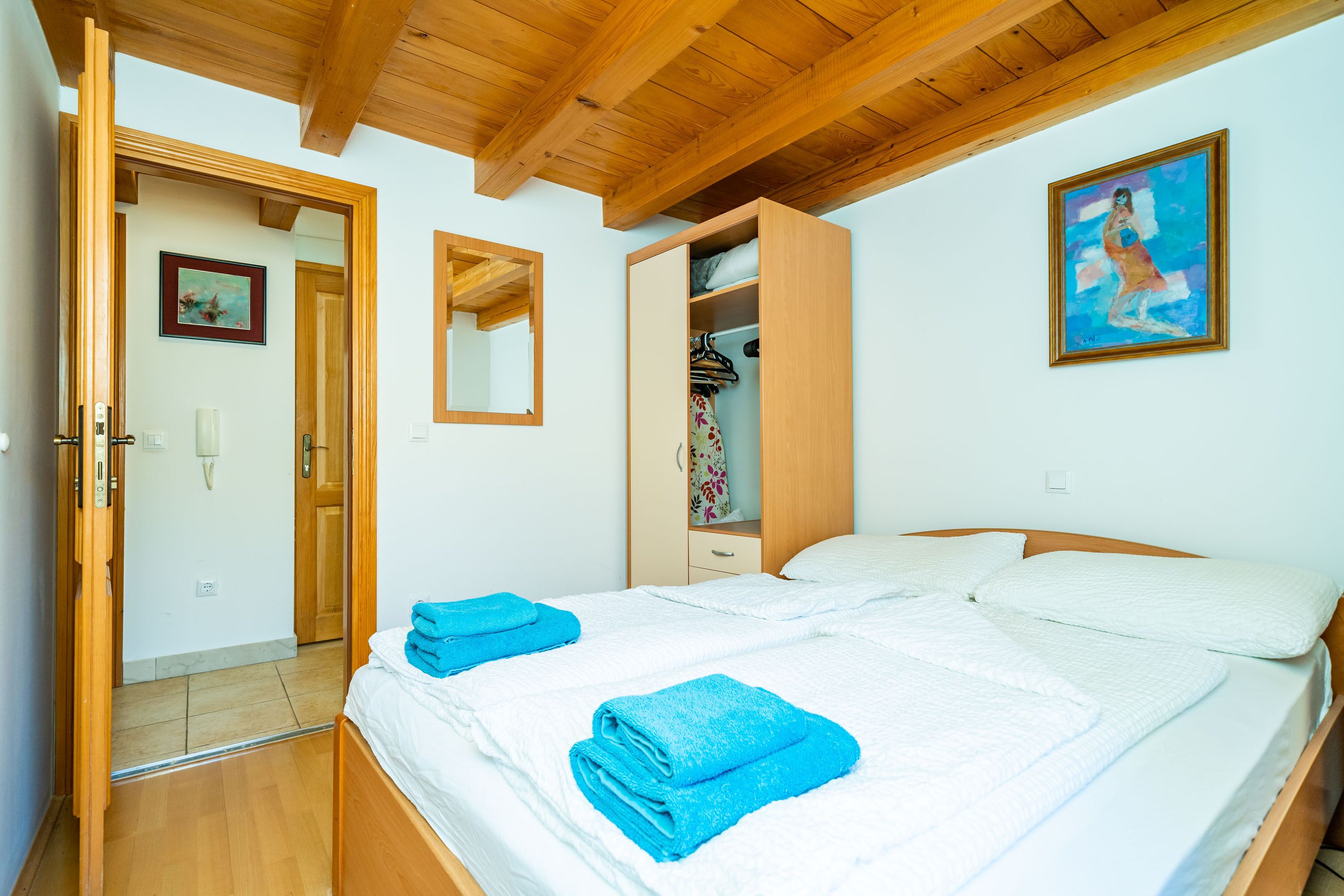 A bedroom with a wooden ceiling, a bed with white bedding and blue towels, a wardrobe, and a painting on the wall.