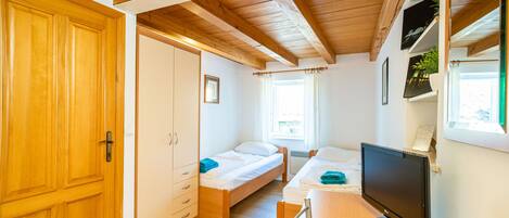 A room with two beds, a desk with a computer, wooden ceiling beams, and a large window.