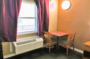 Una habitación con ventana, cortinas moradas, una mesa con dos sillas y un espejo redondo sujeto a la pared.