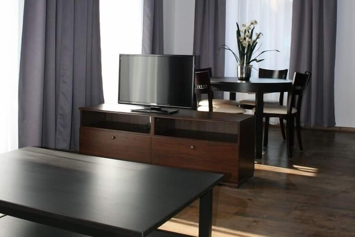 Una habitación moderna con televisión sobre una mesa de madera, una zona de comedor con sillas y un florero con flores.