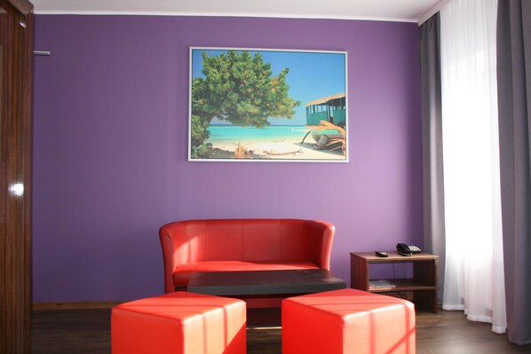 A room with a purple wall, a red sofa, two red ottomans, a small wooden side table with a phone, and a painting of a beach scene.