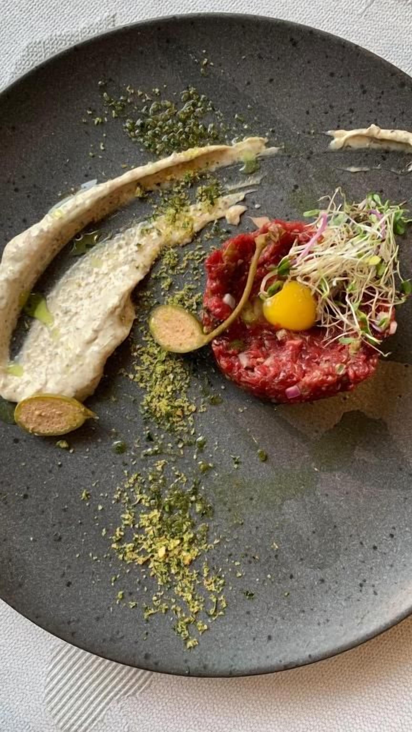 A plate with a dish of raw meat, possibly tartare, garnished with a yellow egg yolk and sprouts, accompanied by slices of cucumber and a green herb.