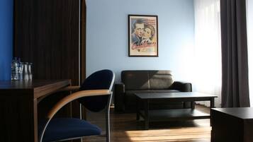 Een kamer met een houten bureau, een blauwe stoel, een zwarte bank en een salontafel. Er hangt een poster aan de muur en er is een raam met gordijnen.