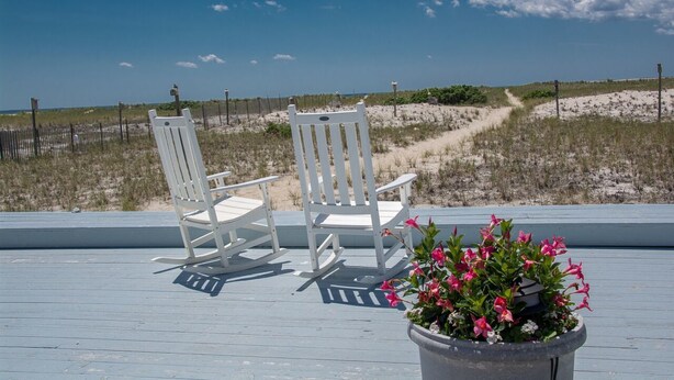 A deck with two white rocking chairs and a potted plant with pink flowers. - East Harbour Motel and Cottages (North Truro)
