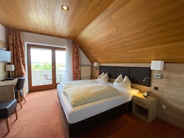 Quarto de hotel com uma cama grande, uma escrivaninha com cadeira, uma televisão e uma varanda com cortinas.
