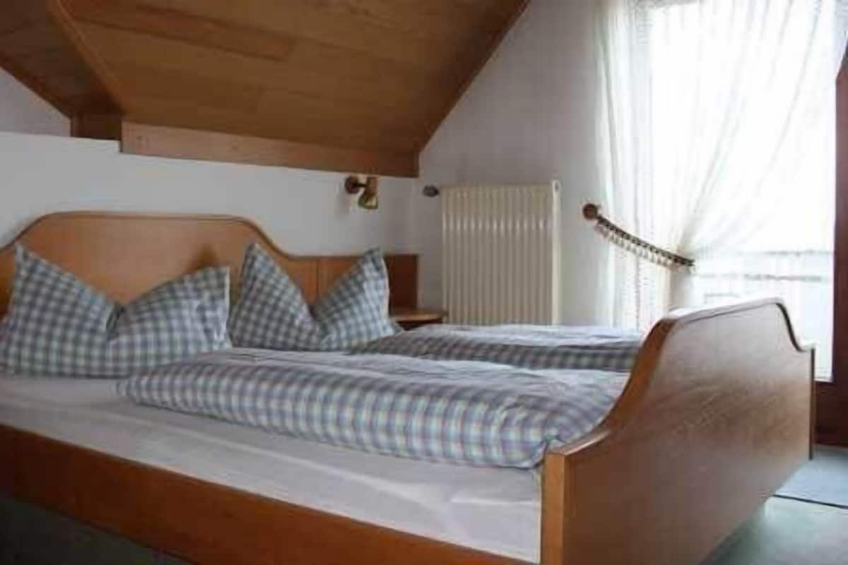 A double bed with a wooden headboard and a checkered bedspread. There is a nightstand with a lamp and a telephone on it. A radiator is visible in the background.