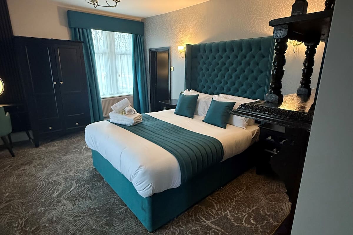 A neatly made bed with a teal headboard, a dark wooden nightstand, and a wall-mounted lamp.