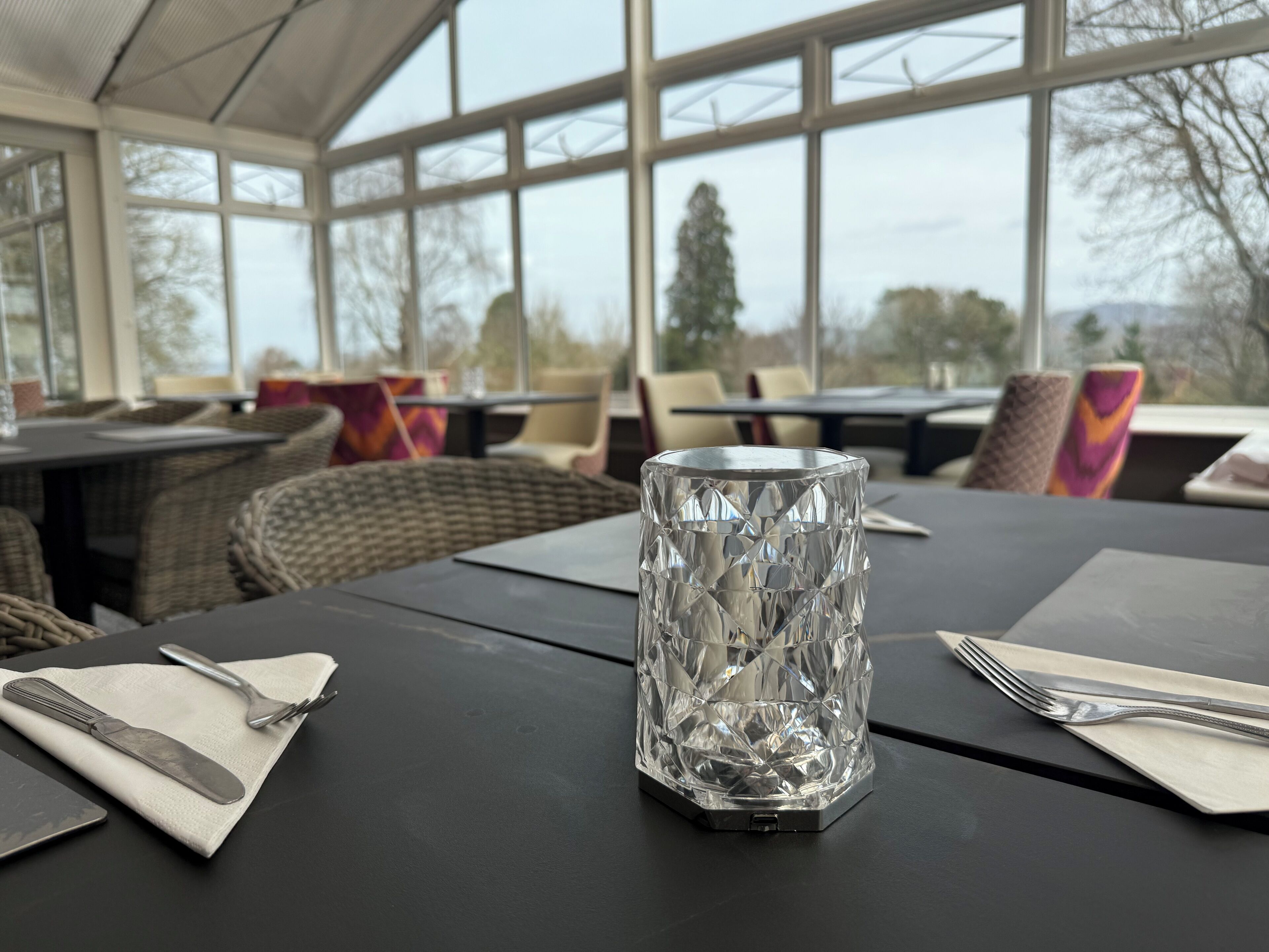 A table with a clear glass centerpiece, a napkin, and cutlery. The setting includes wicker chairs and a glass-enclosed dining area with large windows.