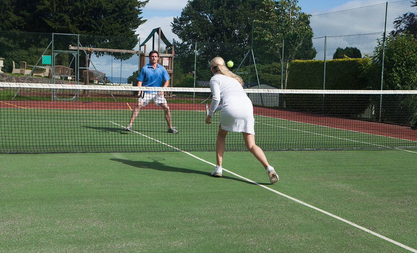 Tenniskenttä, jossa on verkko, kaksi pelaajaa ja peliä varten käytettävä tennispallo.