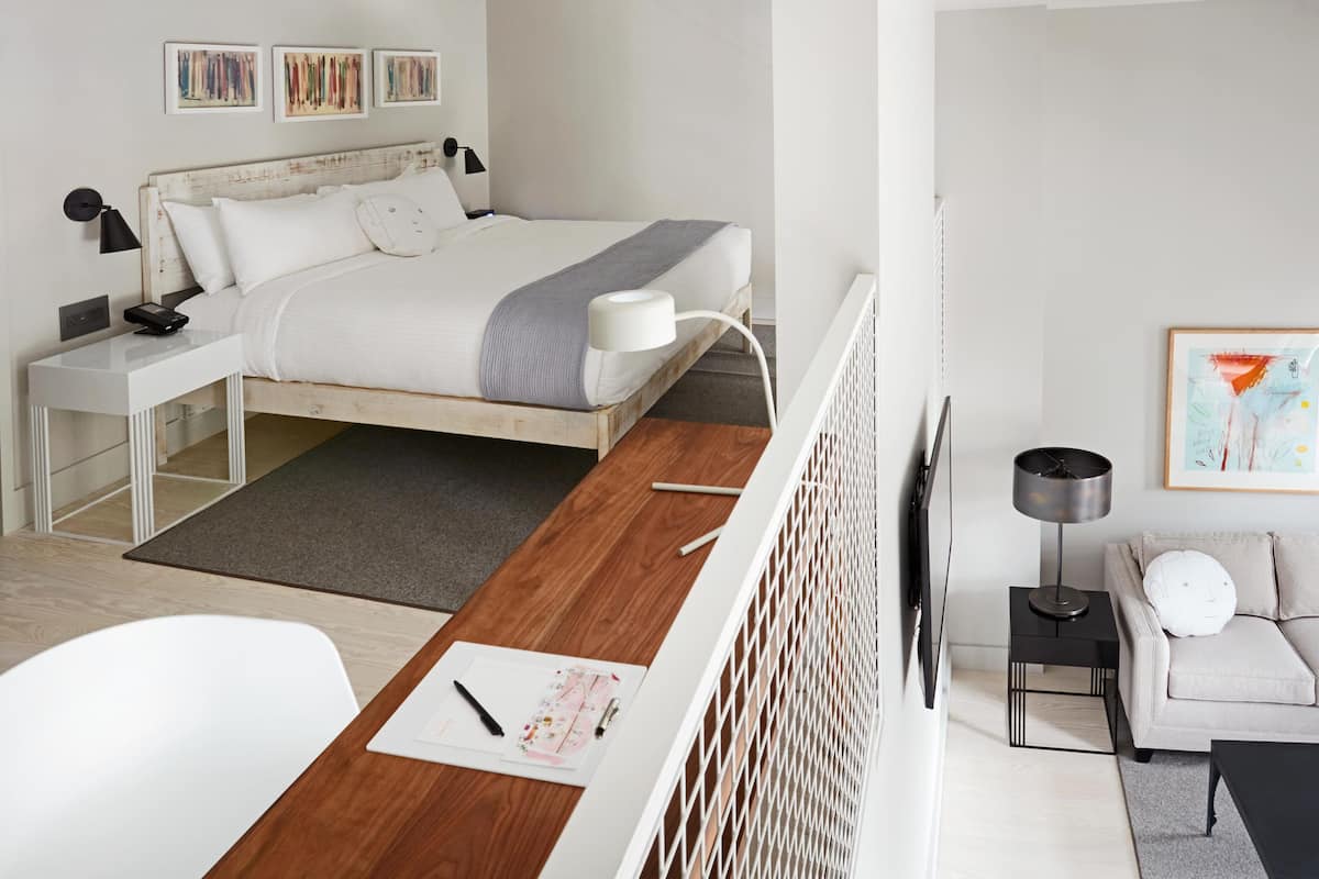 A modern bedroom with a wooden bed, white walls, and a desk with a computer and papers.
