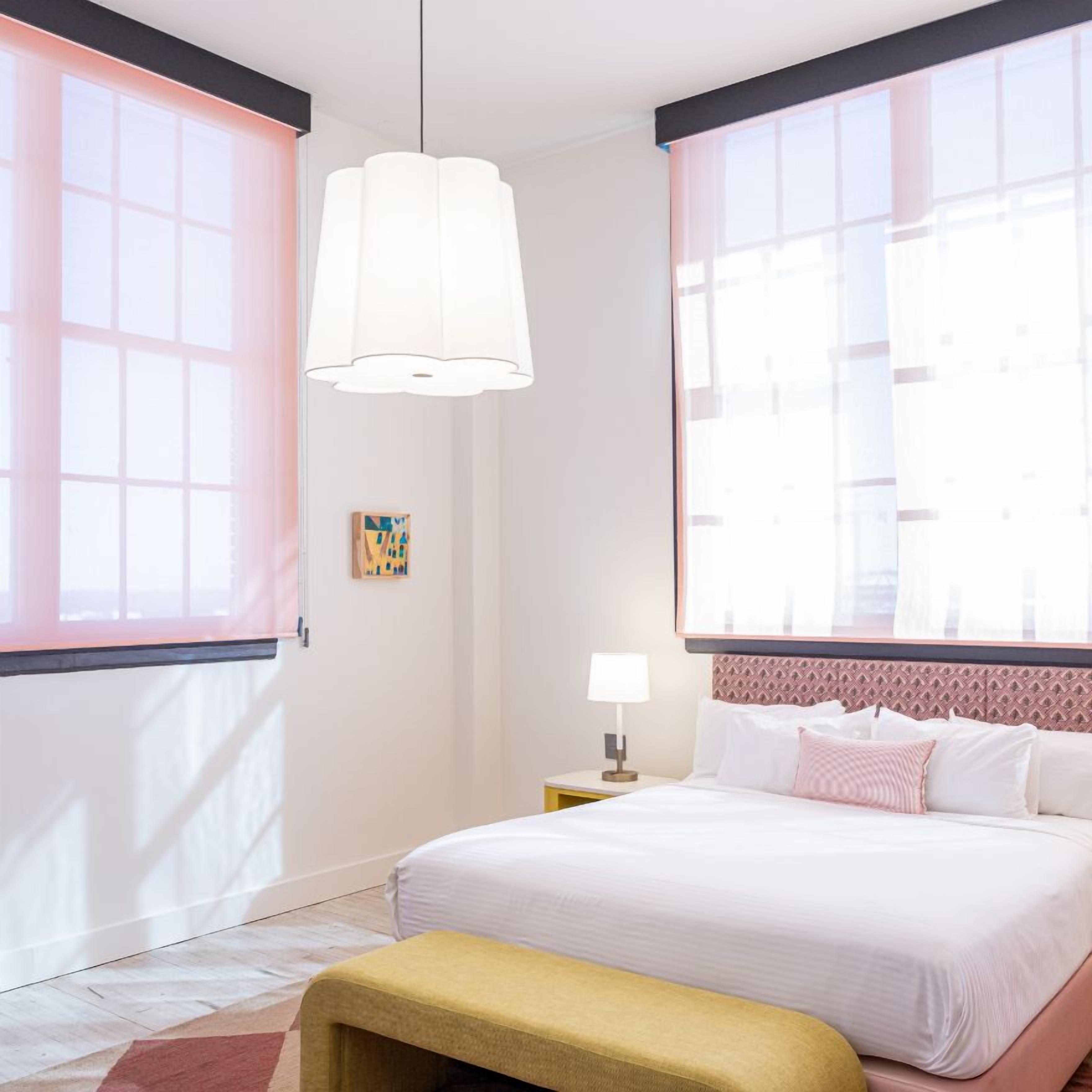 A hotel room with a bed, a bench, a nightstand, a lamp, and a window with curtains.