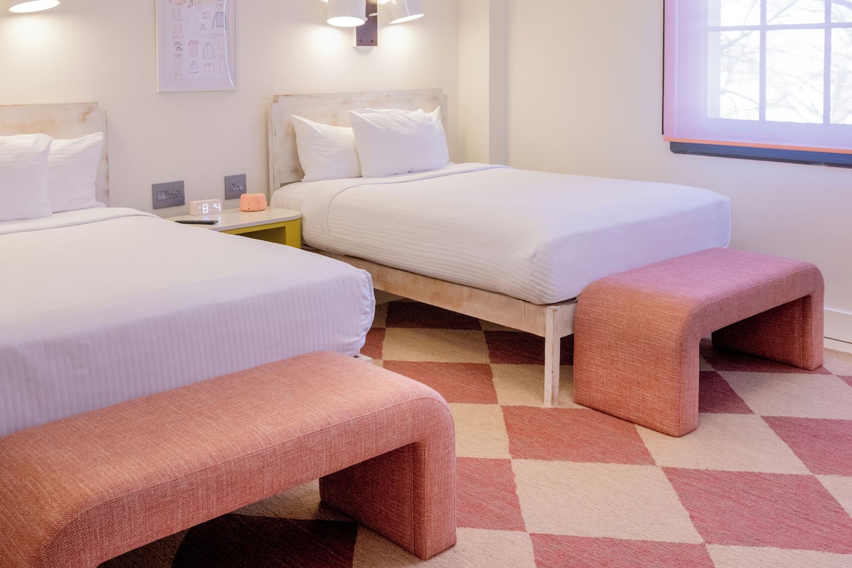 A hotel room with two beds, a bench, a clock, and a window with a view.