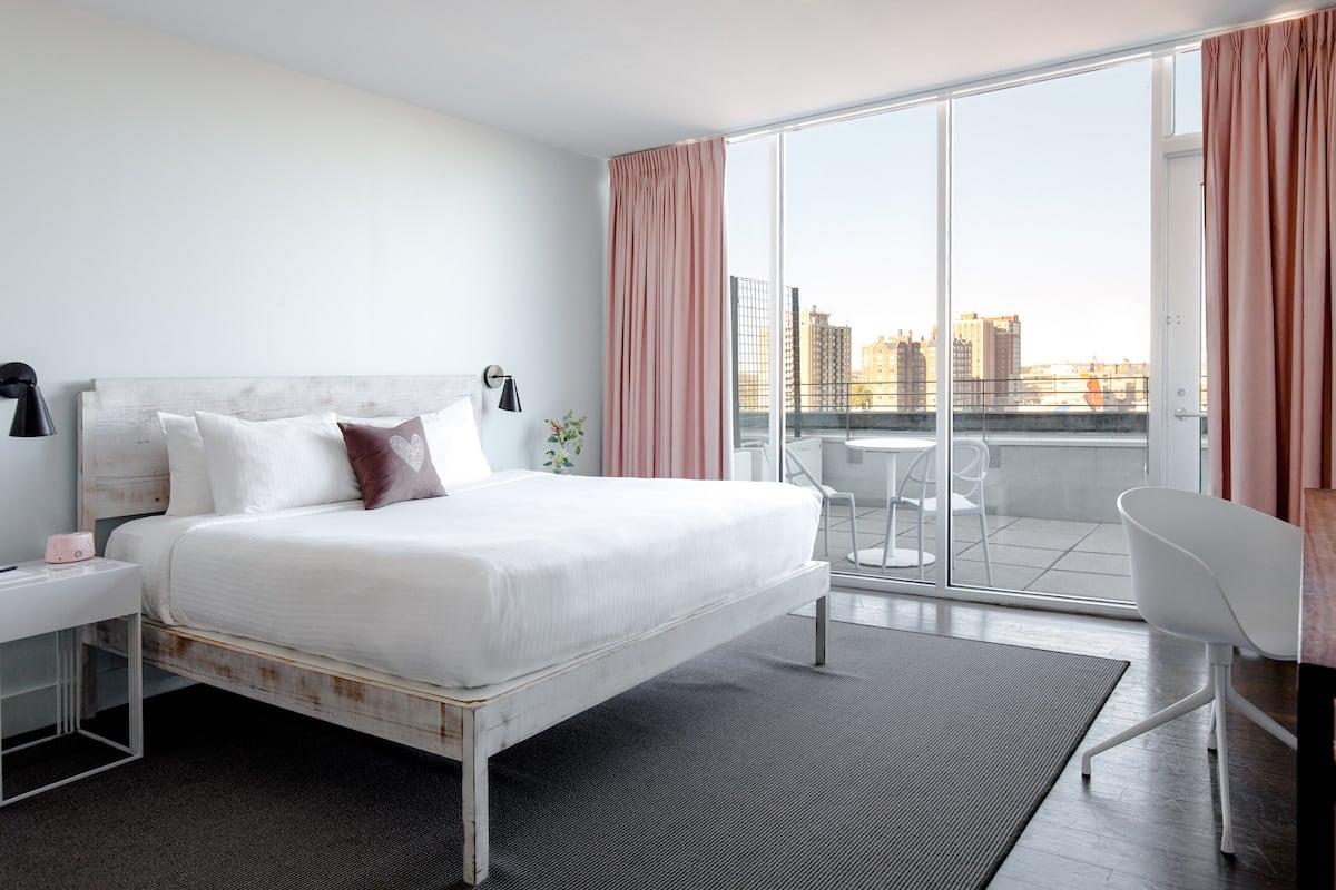 A modern hotel room with a large bed, a desk, a chair, and a balcony with a city view.