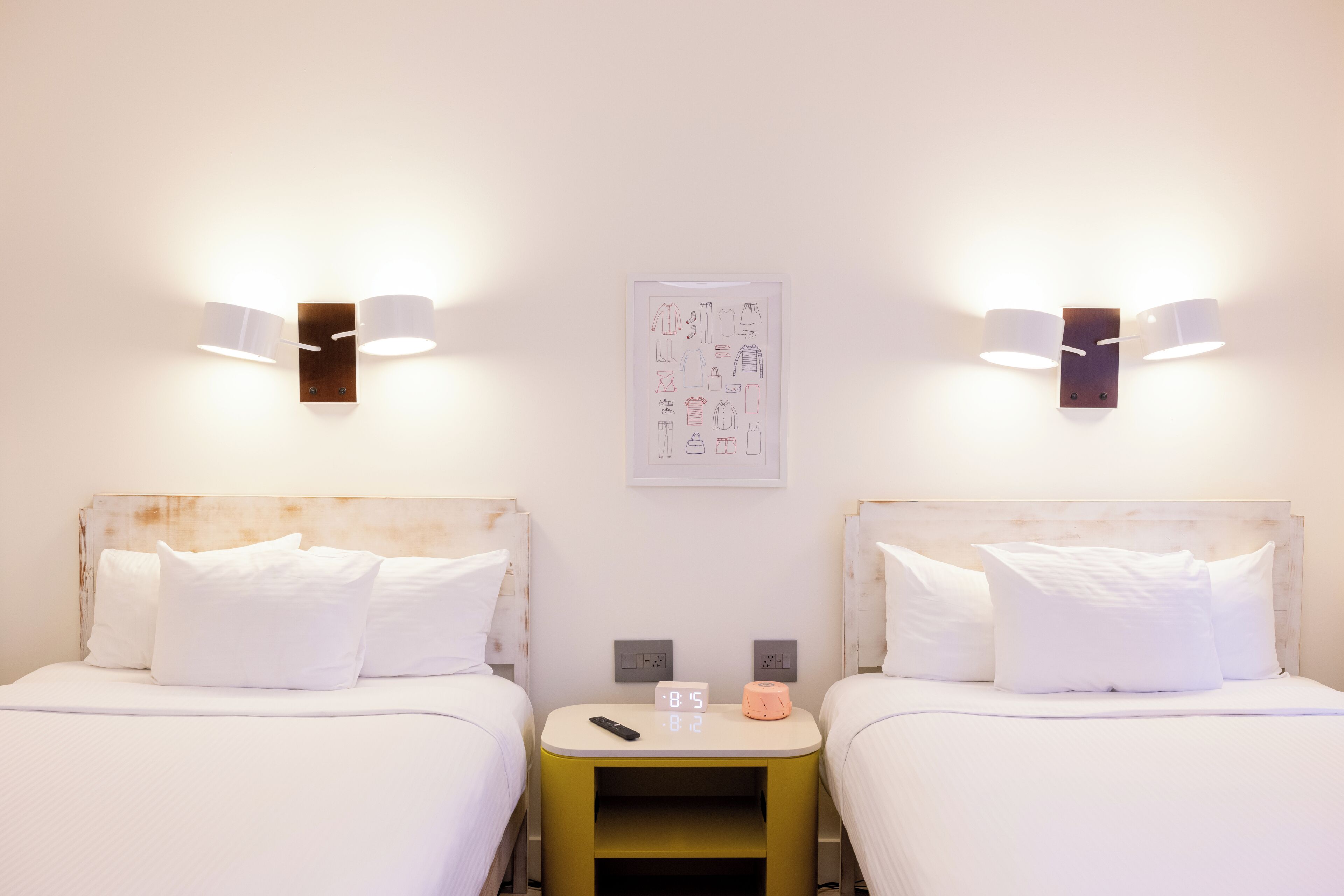 Hotellihuone, jossa on kaksi sänkyä, yöpöytä, jossa on kello, ja seinään kiinnitetyt lamput.