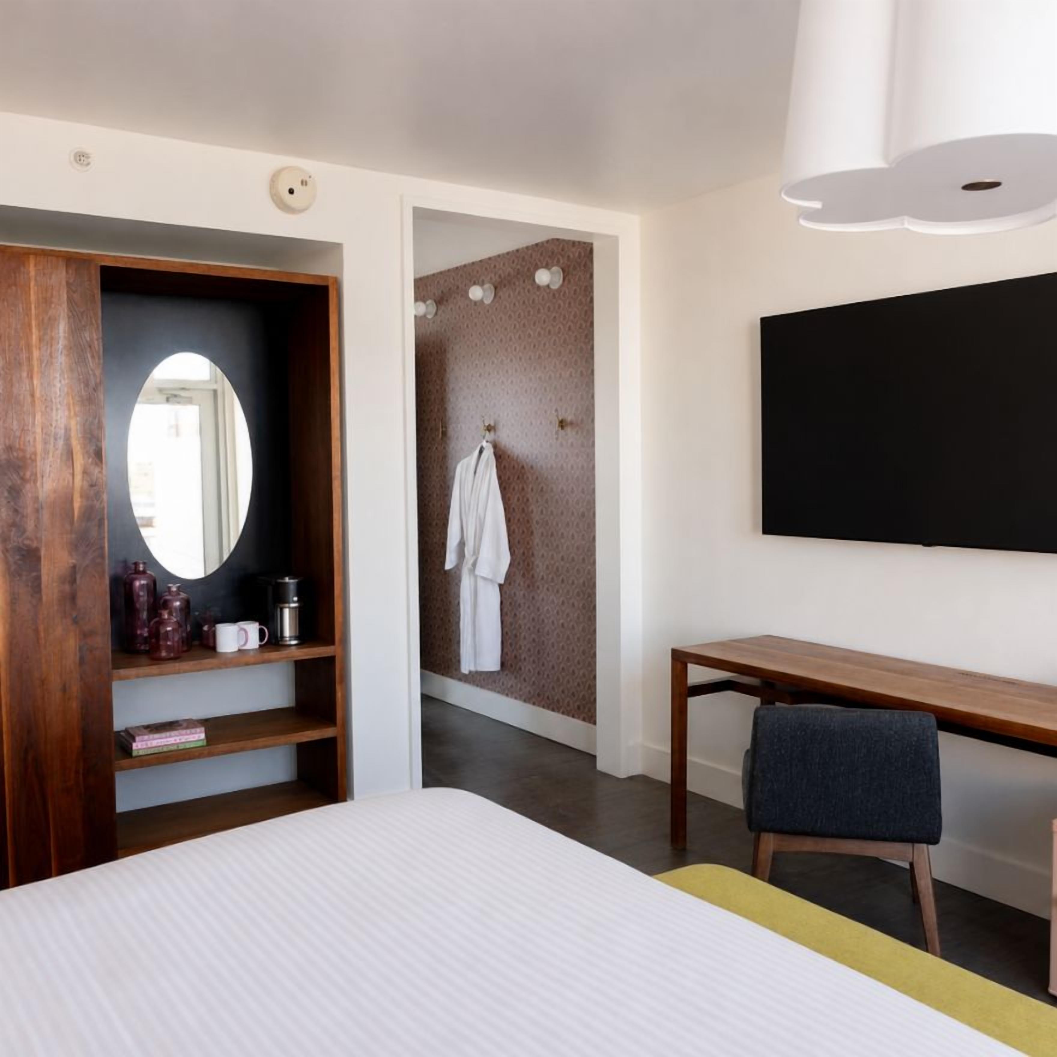A hotel room with a bed, a desk, a chair, a wardrobe, a mirror, a towel rack, and a television.