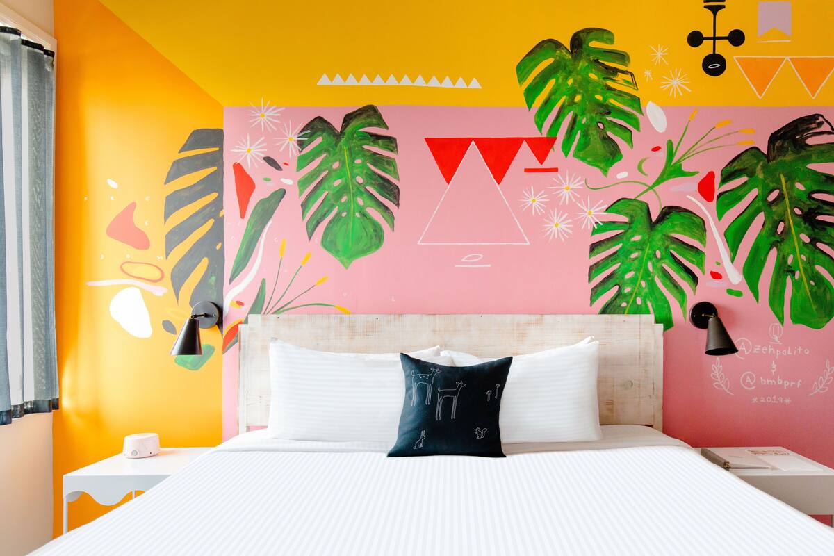 A bedroom with a bed, white bedding, a nightstand, a lamp, and a wall with tropical leaf and geometric patterns.