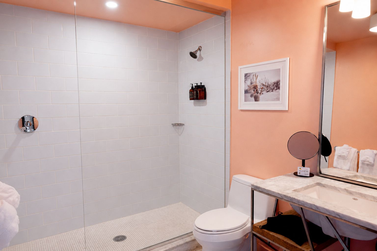 A bathroom with a white tiled shower, a toilet, a sink with a mirror, and a framed picture on the wall.