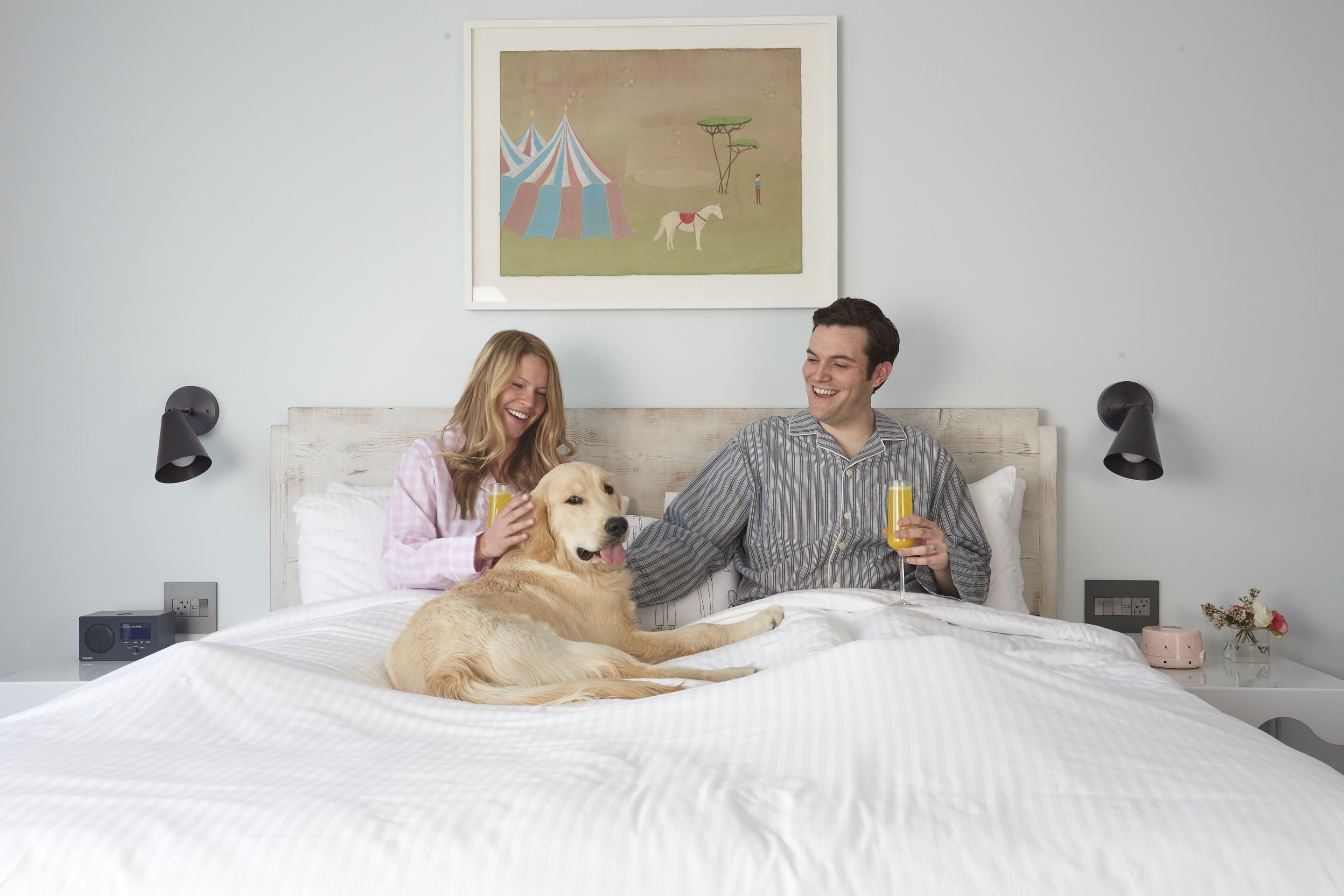 A bedroom with a bed, a dog, two people, a painting, bedside lamps, a clock, and a nightstand with a vase and flowers.