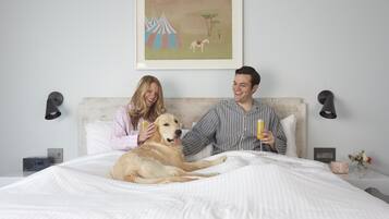 A bedroom with a bed, a dog, two people, a painting, bedside lamps, a clock, and a nightstand with a vase and flowers.