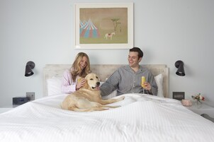 Un dormitorio con cama, un perro, dos personas, un cuadro, lámparas de noche, un reloj y una mesita de noche con un jarrón y flores.