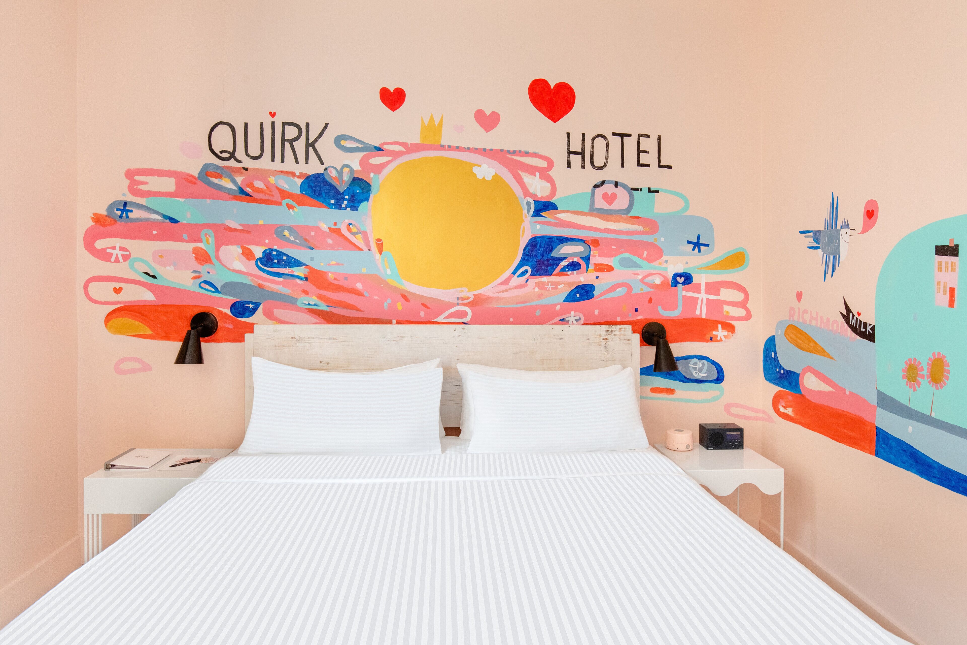 A bed with white bedding, a nightstand with a lamp, and a wall with a colorful abstract design and the words "QUIRK HOTEL".