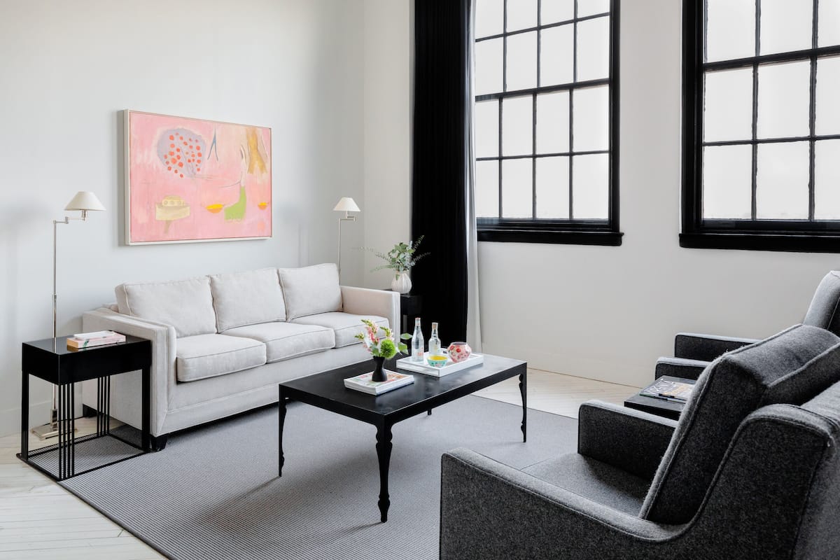 A modern living room with a sofa, a coffee table, and a side table. There is a painting on the wall and a window with black frames.