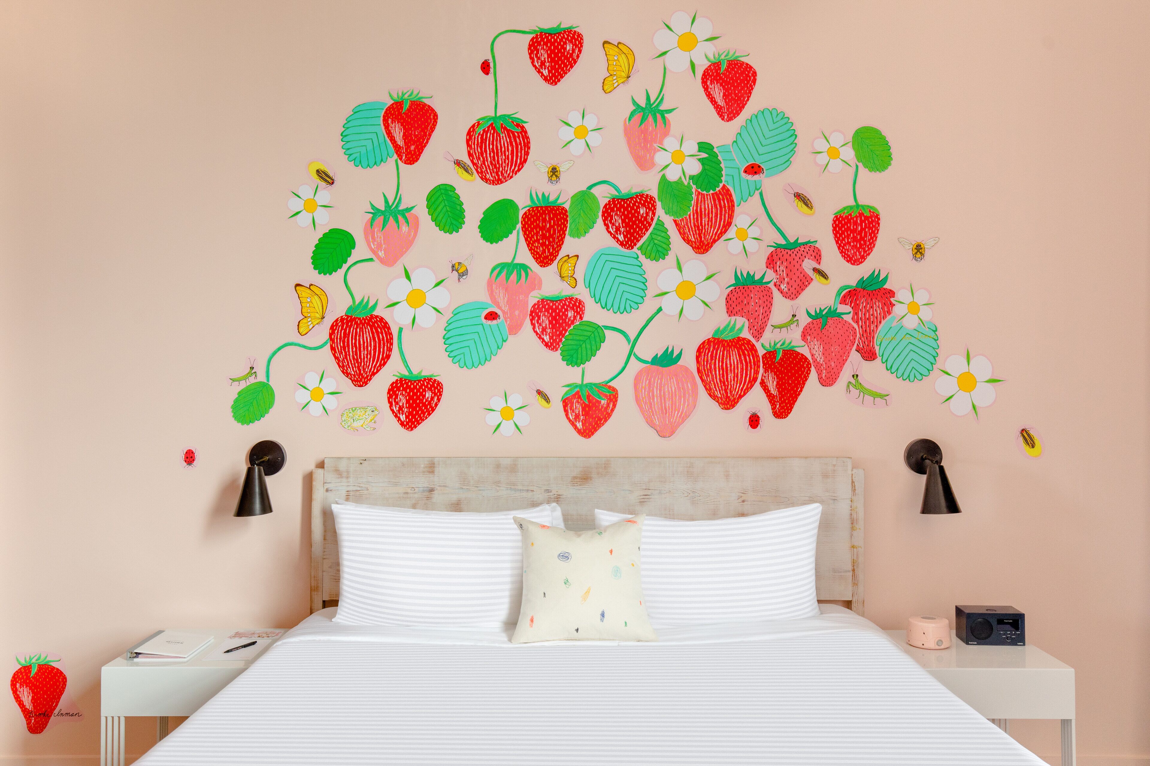 A bedroom with a bed, white bedding, a bedside table with a lamp, a nightstand, and a wall decal of strawberries and flowers.