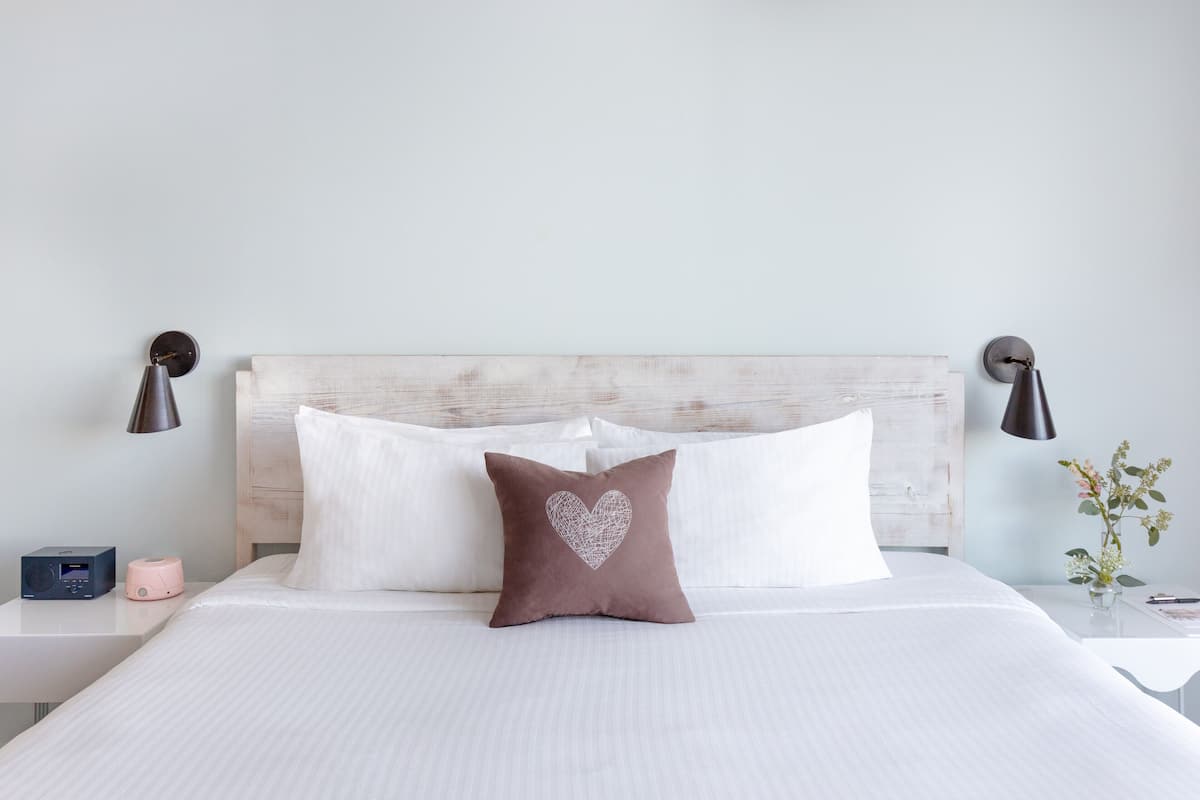 A neatly made bed with white linens, a brown heart-patterned pillow, and two bedside lamps.
