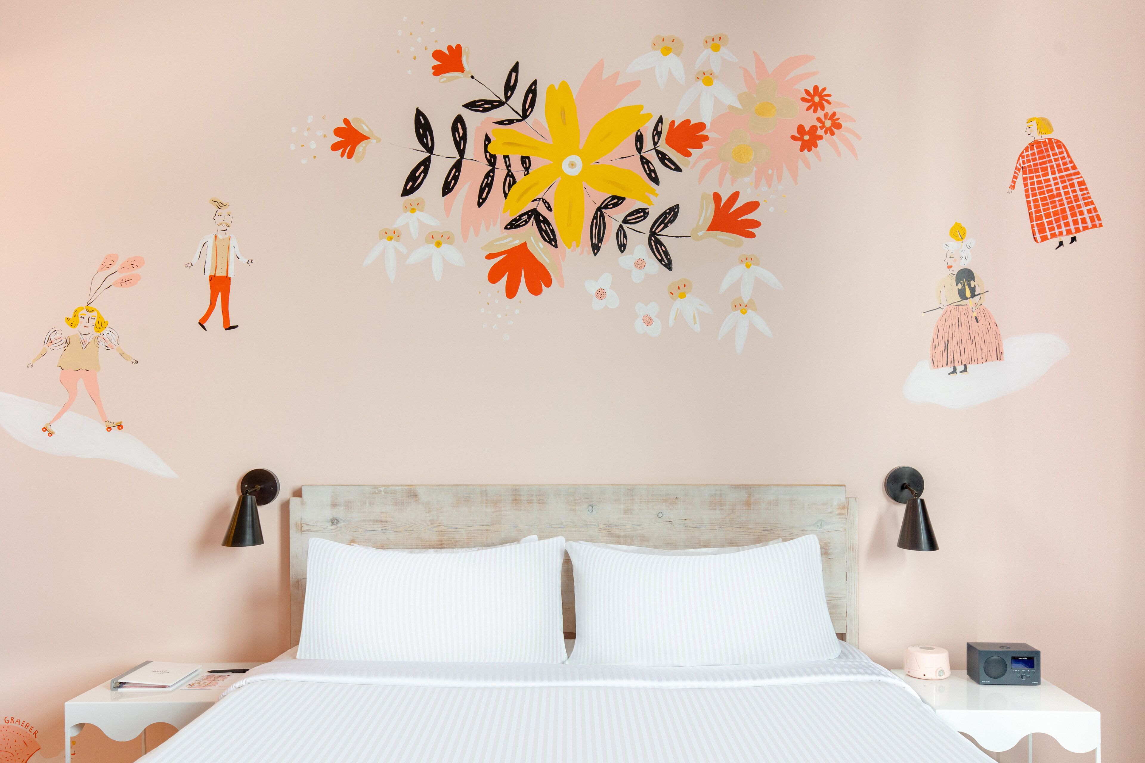 A bedroom with a bed, white bedding, a nightstand with a lamp, and a wall with floral and character decals.