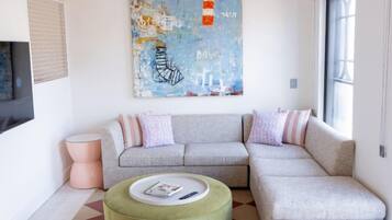 Un salon moderne avec un canapé d’angle, un pouf rond et un tableau abstrait aux couleurs vives.