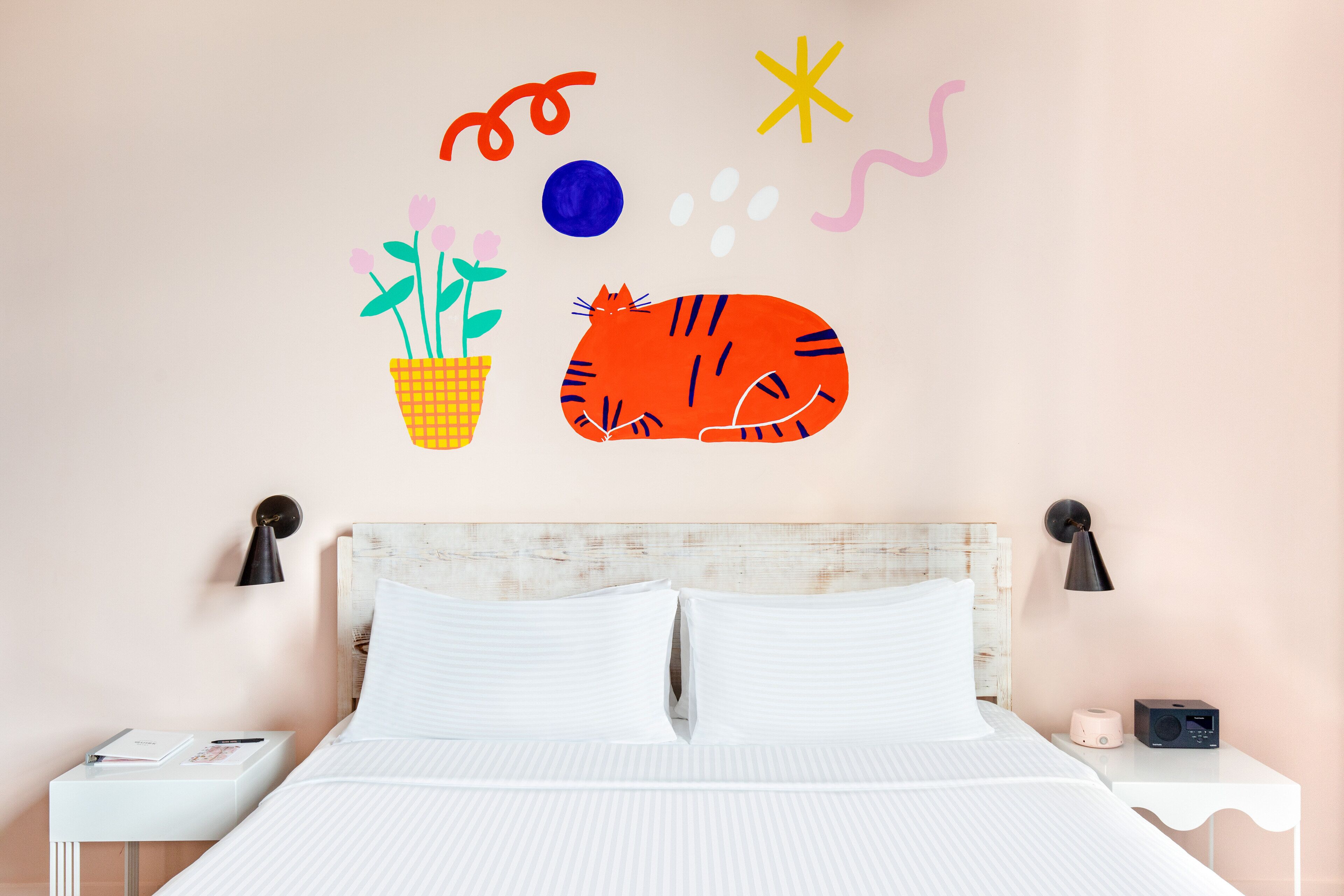Une chambre à coucher avec un lit, du linge de lit blanc, une table de chevet avec une lampe, une horloge murale et un motif décoratif mural représentant un chat, des fleurs et des formes abstraites.