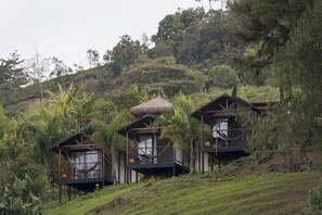 A hillside with three wooden cabins, each with a thatched roof and surrounded by lush vegetation. - Hotel Pietrasanta (El Peñol)