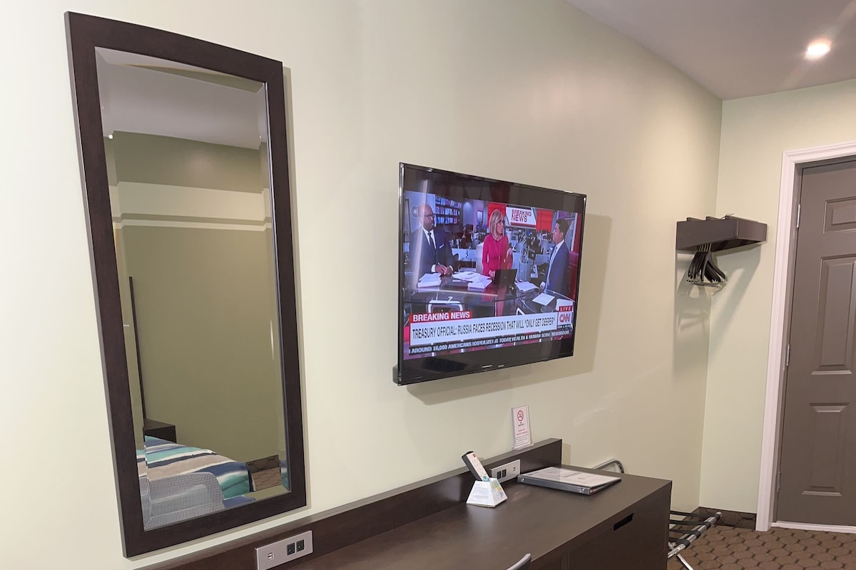 A hotel room with a dark wooden desk, a large mirror, a television displaying news, and a bed with a striped blanket.