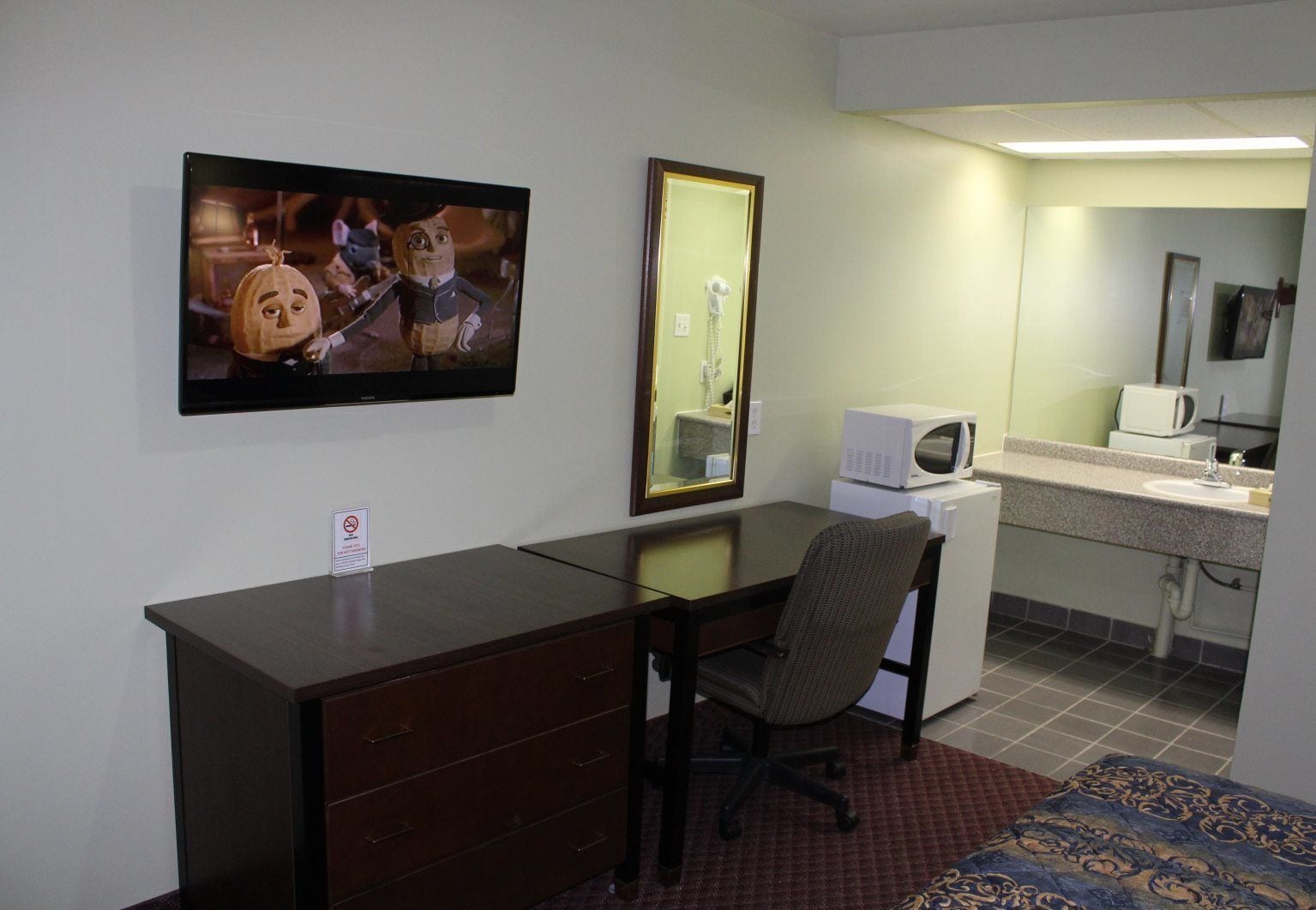 Een hotelkamer met een bureau, stoel, een aan de muur bevestigde televisie, een magnetron en een klein kitchenette.