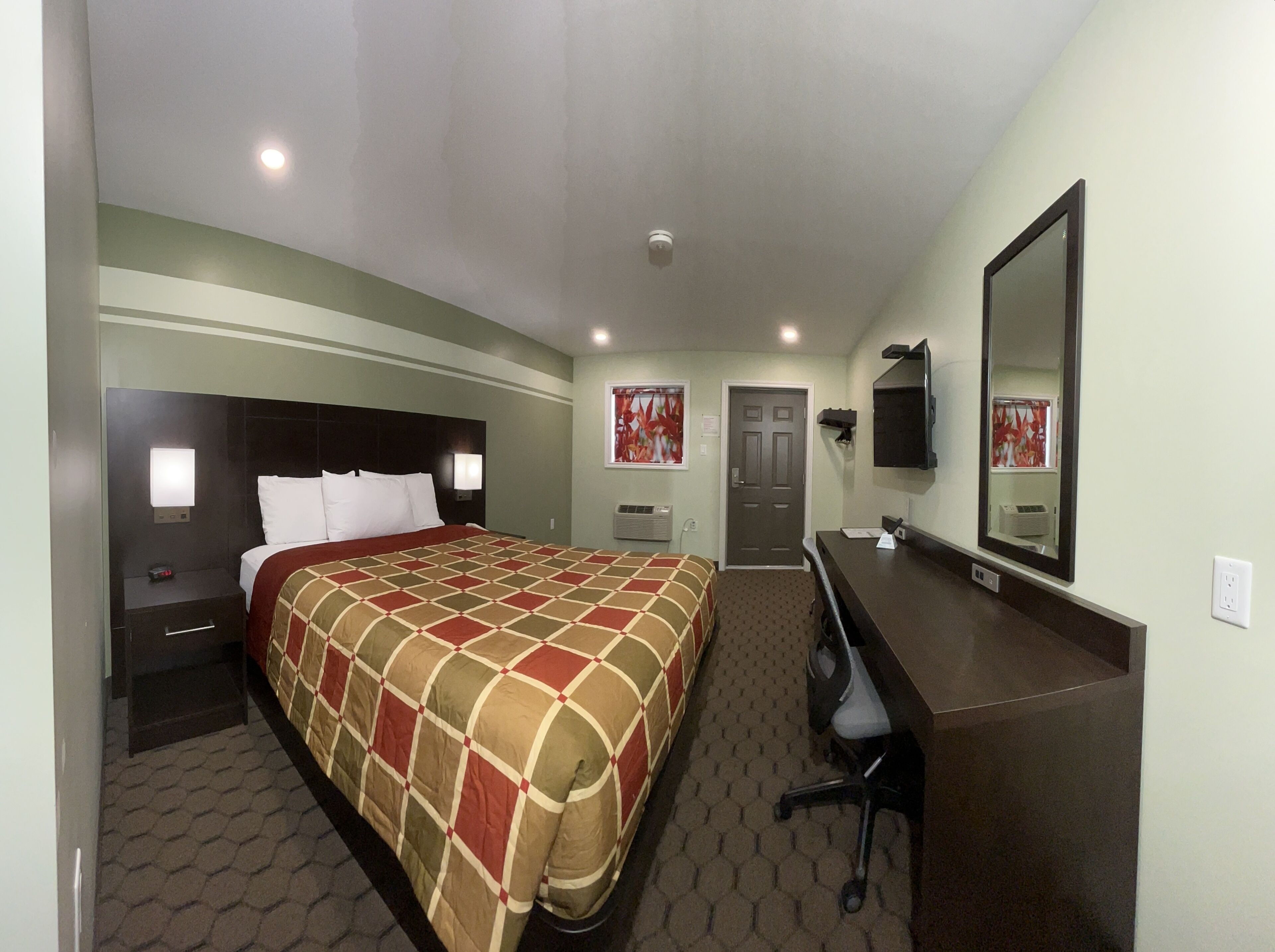 A hotel room with a large bed, a desk, a chair, a mirror, and a door.